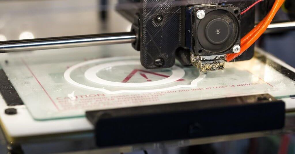 Detailed image of a modern 3D printer producing a polymer firearm lower receiver on a workbench with tools and safety equipment in the background