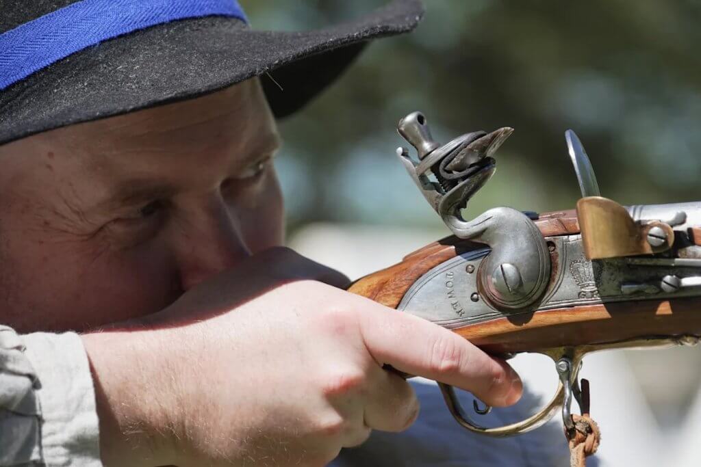 Reenactor firing a reproduction 18th-century musket at an outdoor range, with smoke billowing from the muzzle amid a group of historical enthusiasts.