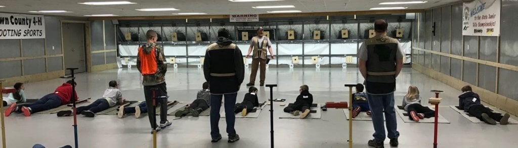 Group of young 4-H participants in safety gear practicing rifle shooting on an outdoor range under adult supervision